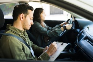 A young couple in a car, the man filling out a form while the woman driving the car